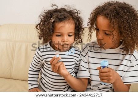 A tender sibling mixed race scene as a curly-haired girl blows bubbles for her younger sister, both dressed in matching stripes, used for National Siblings day and sisterhood. High quality photo