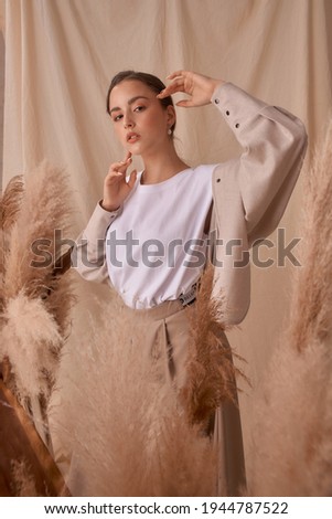 Tender soft studio portrait of elegant female model posing between ears of rye at textile background. Girl in brown skirt and beige jacket. Young woman with brunnette hair in pony tail