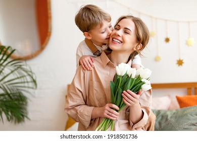 zärtziger Sohn küsst die glückliche Mutter und gibt ihr einen Tulpen-Bouquet, gratuliert ihr zum Muttertag während der Feiertagsfeier zu Hause
