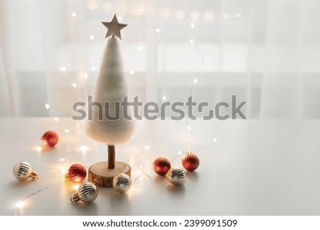 Tender white Chhristmas tree with baubles in a backlight of the window with fairylights. Festive magical still life composition with copy space for your text.