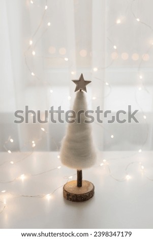 Tender white Chhristmas tree with wooden star in a backlight of the window with fairylights. Festive holidays concept.