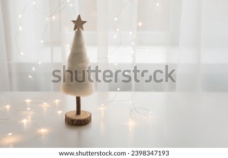 Tender white Chhristmas tree with wooden star in a backlight of the window with fairylights. Festive holidays conbcept, copy space for your text.