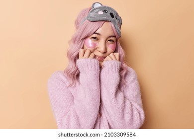 Tender Young Asian Woman Keeps Hands Under Chin Applies Hydrogel Patches For Skin Treatment Wears Sleepmask And Cashmere Jumper Isolated Over Beige Background. Beauty And Antiaging Procedures