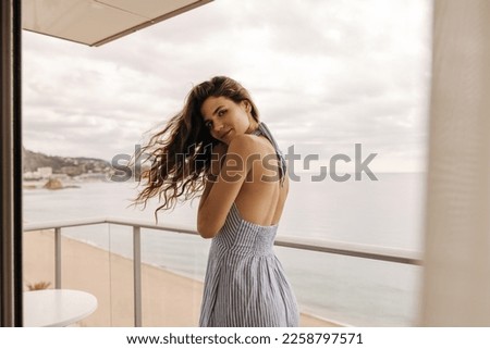 Tender young caucasian girl with wavy hair stands on hotel balcony looking at camera. Brunette woman wear striped dress folded her arms nicely. Concept pleasant stay
