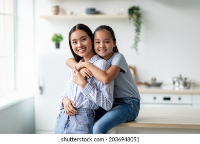 Tenderness And Motherhood Concept. Cute Asian Girl Hugging Young Mother From The Back, Sitting On Dinner Table At Kitchen Interior. Mom And Her Child Spending Time Together At Home