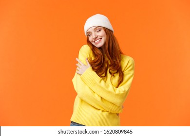 Tenderness, winter holidays concept. Charming romantic redhead woman in soft sweater, white beanie, close eyes and embrace own body, hugging herself, smiling cheerful, orange background