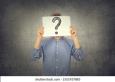 Tennager Boy using a white paper with drawn question mark on gray wall background. Question mark, symbol. Pensive male. Getting answers. Doubtful man holding Question Mark. Problems and solutions. 