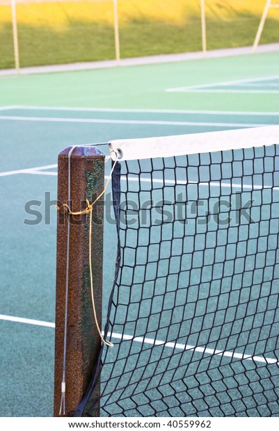 Tennis court net attached to rusting metal post\
with yellow and grey ropes.  Tennis court of green tarmac is\
visible.  Out of focus wire fence and shadowed grassy hill in\
background.  Winter\
daylight.