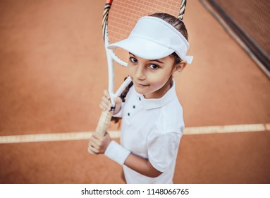 Tennis Is My Favorite Game! Portrait Of A Pretty Sporty Child With A Tennis Racket. Little Girl Smiling At Camera On Tennis Court. Training For Young Kid.