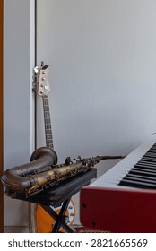 A tenor saxophone on its case, a four-string bass guitar and a red electronic keyboard share a compact home music corner. The sparse interior arrangement leaves a broad plain wall above the grouped instruments. The uncluttered arrangement suits themes of personal practice and small ensemble hobbies.