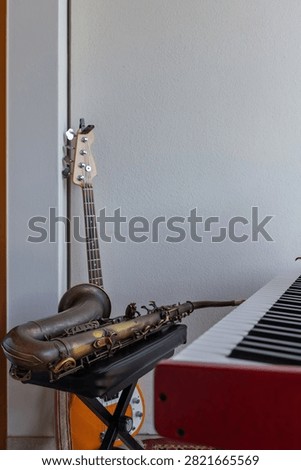 A tenor saxophone on its case, a four-string bass guitar and a red electronic keyboard share a compact home music corner. The sparse interior arrangement leaves a broad plain wall above the grouped instruments. The uncluttered arrangement suits themes of personal practice and small ensemble hobbies.