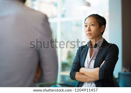 Tense Asian businesswoman looking at male partner with crossed arms. Two colleagues confronting each other in office space. Clashing personalities concept
