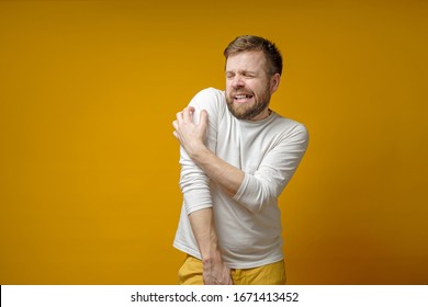 Tense Bearded Man Scratches The Itchy Skin On Arm, Through Clothes, Painfully Grins Teeth And Squints Eyes. Health Concept.