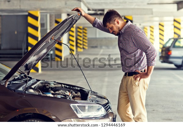 A tense and frustrated young man\
driver stands next to a broken car and looks under the hood in the\
Parking lot . Problems with travel and the concept of\
care.