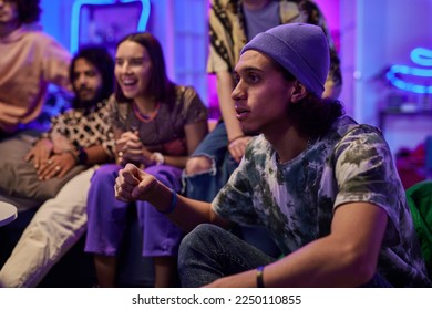 Tense guy in beanie hat and t-shirt looking at tv screen during match broadcast against group of happy multicultural friends - Powered by Shutterstock