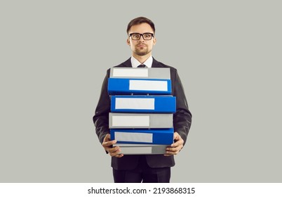 Tense Male Office Worker With Stressed Expression Holding Stack Of Heavy Folders On Gray Background. Overworked Young Man In Suit And Glasses Suffering From Overloaded Work. Banner. Isolated.