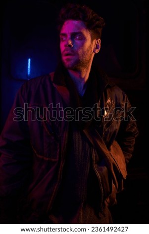 tense man in black jacket looking away in darkness of abandoned underground tunnel with neon light