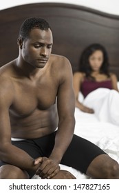 Tensed Shirtless African American Man Sitting On Bed With Blurred Woman In Background