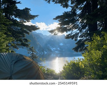 A tent is set up by a mountain lake, framed by evergreen trees. Sunlight filters through the trees, casting reflections on the water with snow-capped peaks in the background. - Powered by Shutterstock