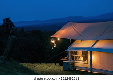 A tent is set up in a grassy field at night. The tent is white and has a tarp over it. The tent is lit up with lights, giving it a cozy and inviting atmosphere