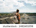 Teotihuacan, Mexico. A Mexican American tourist with a backpack and hat enjoys the view from the top of the Moon Pyramid. The Sun Pyramid and Avenue of the Dead can be seen in the distance.