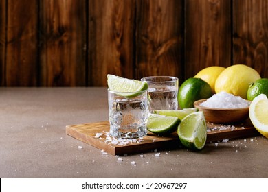 Tequila in a glass served with lemons, limes and salt over brown texture background