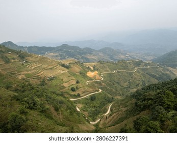 Terraced rice fields cascading down mountain slopes in Ha Giang Loop, Vietnam, with winding dirt roads cutting through agricultural hillsides and misty peaks in the distance.