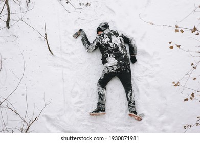 A terribly drunk homeless and dirty man in a black coat lies sleeping on his stomach on white snow in a frosty winter with a bottle of alcohol, strong whiskey. Photography, copy space, concept.