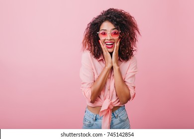 Terrific african model expressing surprised emotions while posing on indoor photoshoot. Stylish curly woman in sunglasses having fun in pink studio.