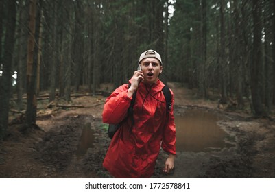 The Terrified Male Traveler Is Talking On The Phone, Shouting In The Mountain Fir Forest. A Young Tourist Man In A Red Jacket With A Backpack Is Feeling Scared And Lost In The Woods.