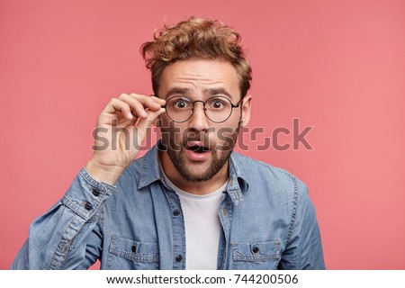 Terrified man dressed casually, stares through glasses, being surprised find out about his success. Male doesn`t believe his own luck, isolated over pink background. Emotions, people concept
