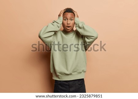 Terrified shocked black young man keeps hands on head has widely opened mouth cannot believe in something impressive dressed in casual jumper ans black trousers isolated over brown background.