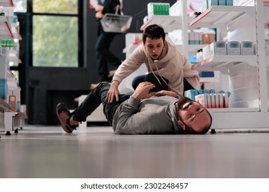 Terrified woman noticing man on drugstore floor having epileptic seizure, running to give first aid for fainting. Apothecary client helping person suffering from epilepsy disorder attack - Powered by Shutterstock