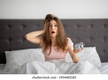 Terrified yound woman holding alarm clock in bed at home, grabbing her head, oversleeping to work. Shocked millennial lady being late, having difficulty getting up in mornings