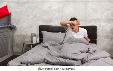 Terrified Young Man Woke Up From A Nightmare And Wiping Sweat From His Forehead While Sitting In Stylish Bed With Grey Colors In A Bedroom In Loft Style