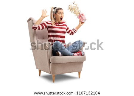 Terrified young woman with a box of popcorn sitting in an armchair isolated on white background
