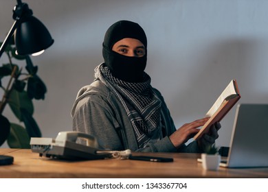 Terrorist In Black Mask Sitting At Table And Reading Book