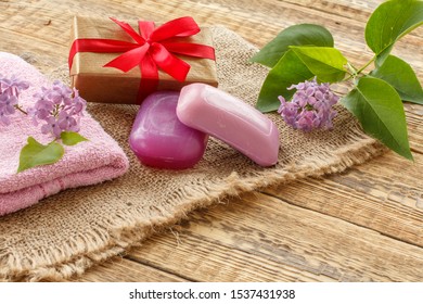 Terry Towel, Soap For Bathroom Procedures, Gift Box And Lilac Flowers On Sackcloth And Old Wooden Boards. Spa Products And Accessories.