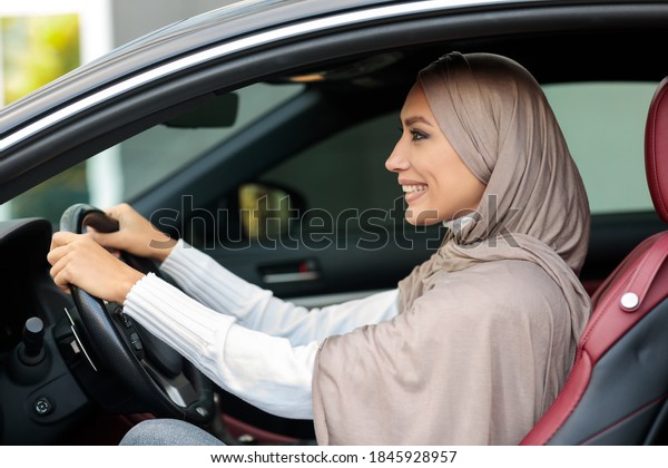 Test Drive Concept. Portrait of smiling muslim\
woman in hijab driving new luxury car, looking at road. Cheerful\
arabian lady in headscarf sitting in salon on front seat, holding\
steering wheel