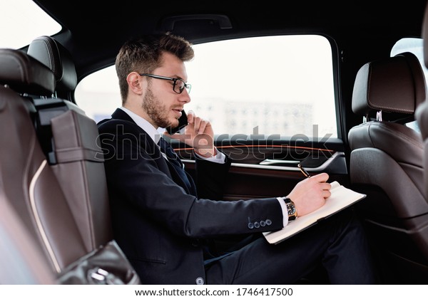 Test Drive\
Concept. Young manager in glasses writing in notebook with pen\
sitting in back seat of new car, side\
view