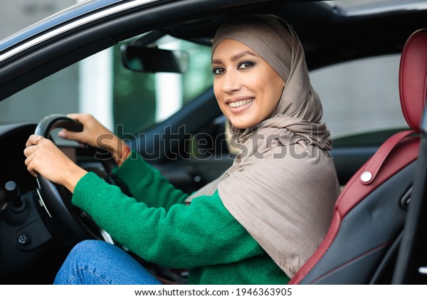 Test Drive. Smiling muslim woman in hijab driving\
new luxury car. Cheerful arabian lady in headscarf sitting in salon\
on front seat with open door, holding steering wheel, looking and\
posing at camera