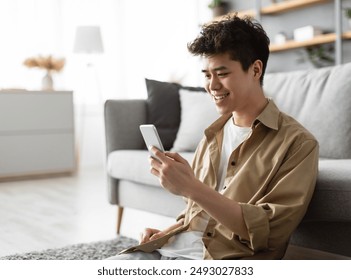 Testing New App. Handsome Asian Man Using Smartphone While Relaxing Leaning On Couch At Home, Happy Guy Sitting On Floor In Living Room, Browsing Internet Or Messaging With Friends, Free Copy Space - Powered by Shutterstock