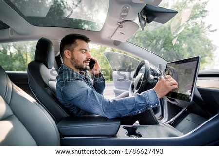 Testing a new electric futuristic car with self driving system. Side view of satisfied Caucasian man in casual jeans shirt sitting in modern car, talking phone and toushing navigation autopilot screen