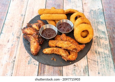 Tex Mex Appetizers With Onion Rings, Cheese Sticks, Chicken Wings, BBQ Wings And Sauces On A Round Black Slate Plate