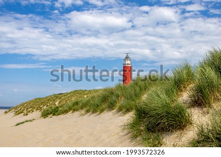 Texel lighthouse in landscape with dunes, grass and beach. On blue sky with clouds. Red light tower in National park 'Duinen van Texel'. Wadden Island Texel, The Netherlands.