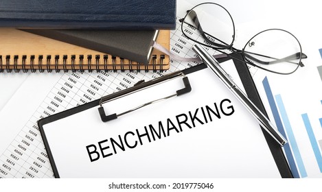 Text BENCHMARKING on Office desk table with notebooks, supplies,analysis chart, on white background.