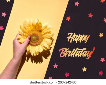 Text Happy Birthday Cut Out Of Orange And Red Paper. Hand Holds Natural Sunflower. Flower In Hand, Small Floral Elements On Blackboard. Diagonal Split Yellow And Black Background. Flat Lay, Top View.