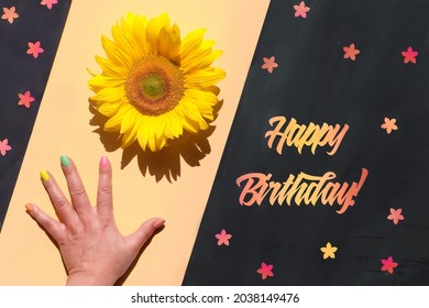 Text Happy Birthday Cut Out Of Orange And Red Paper. Hand Holds Natural Sunflower. Flower In Hand, Small Floral Elements On Blackboard. Diagonal Split Yellow And Black Background. Flat Lay, Top View.