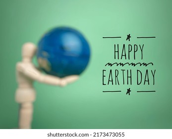 Text Happy Earth Day With Blur Wooden Mannequin Holding Eart Globe On A Blur Green Background.