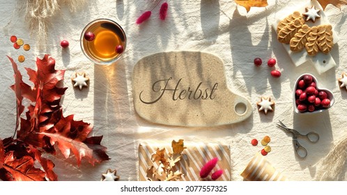Text Herbst means Autumn in German language. Autumn decorations - cranberry, cookies. tea and dry oak leaves. Flat lay, top view on uncolored textile. Fall background, sunlight with long shadows.